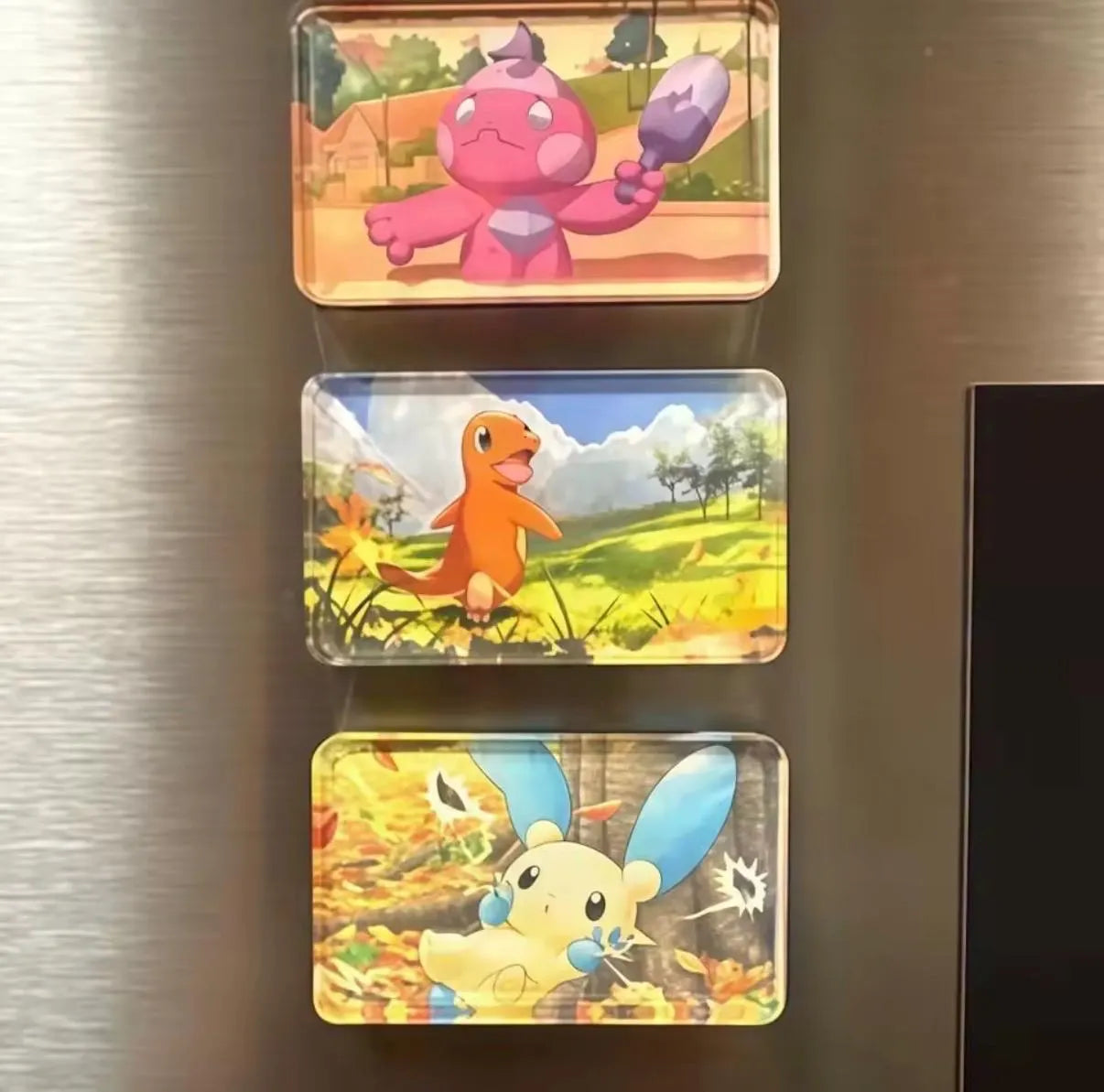 Three rectangular acrylic magnets featuring colorful Pokémon characters (a pink creature, an orange bird, and a cream creature with blue ears) arranged vertically on a brushed stainless steel surface.